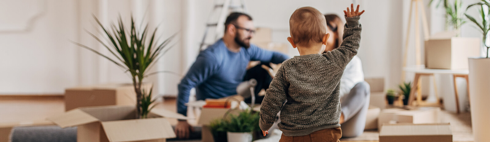 A toddler walks toward parents in the background