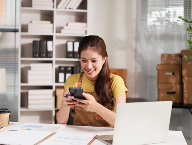 Young Asian woman entrepreneur working at a home office, handling her online business, using a smartphone for communication and order management, with a laptop and documents on the desk